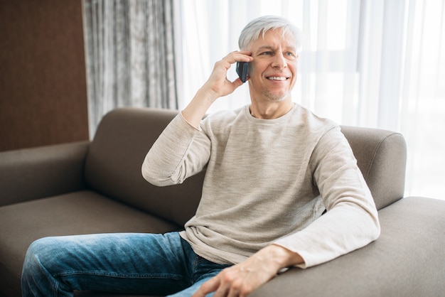 Homme adulte assis sur un canapé à la maison et parler par téléphone mobile. Personne de sexe masculin mature en jeans se détendre dans un fauteuil