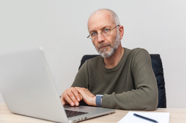 Homme adulte aux cheveux gris travaillant sur ordinateur portable et regardant la caméra