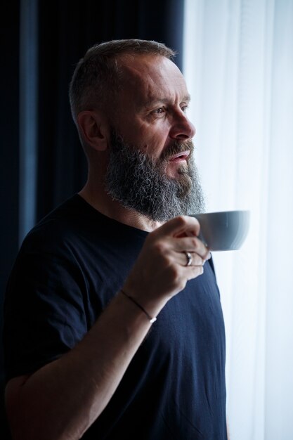 Un homme adulte avec une barbe boit du café et regarde par la fenêtre. Concept de loisirs sur le terrain de la journée de travail