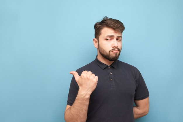 Homme adulte avec une barbe se dresse sur le bleu, regarde pensivement sur le côté et jette son doigt dans un espace vide