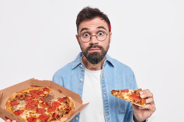 Un homme adulte barbu étonné tient une tranche de pizza mange de la restauration rapide a surpris l'expression une barbe épaisse porte des vêtements décontractés