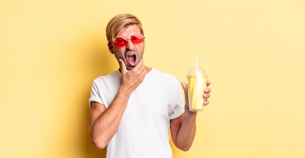 homme adulte blond avec la bouche et les yeux grands ouverts et la main sur le menton avec un milk-shake