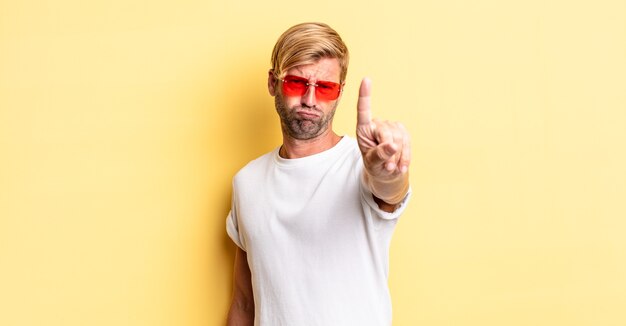 homme adulte blond souriant et à l'air sympathique, montrant le numéro un et portant des lunettes de soleil