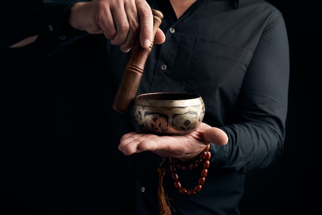 Homme adulte dans une chemise noire tourne un bâton en bois autour d'un bol d'eau tibétain en cuivre