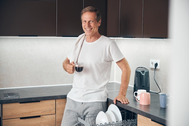 Homme adulte détendu souriant étant dans la cuisine et savourant son café aromatique