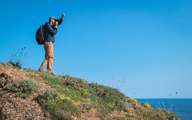 Un homme adulte avec du matériel photographique se tient au bord d'une falaise Ci-dessous se trouve une baie de mer pittoresque avec des bateaux de pêche