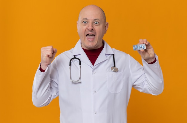 Homme adulte excité en uniforme de médecin avec stéthoscope tenant un blister de médicaments et gardant le poing levé