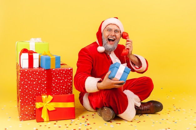 Homme adulte extrêmement excité portant un costume de père Noël assis près de nombreuses boîtes avec des cadeaux de Noël, parlant de voeux avec des vacances via un téléphone fixe, prise de vue en studio intérieur isolé sur fond jaune