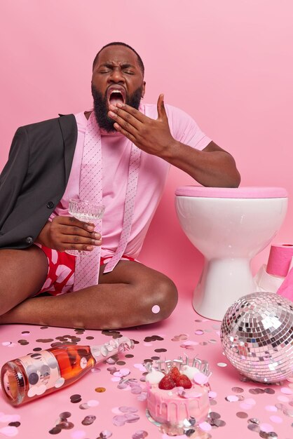 Un homme adulte fatigué à la peau foncée bâille et couvre la bouche avec la main veut dormir après que la fête tient un verre de cocktail se penche sur la cuvette des toilettes pose dans la salle de toilette avec le désordre autour de fond rose