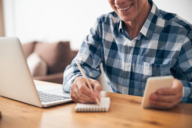 Homme adulte gai assis sur son lieu de travail et prenant des notes lors d'une conférence en ligne, profitant de son processus de travail à la maison