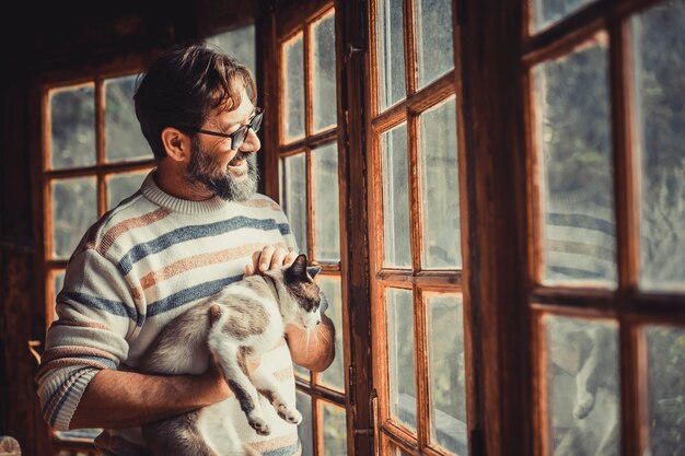 Un homme adulte heureux et mature avec un chat regarde par les fenêtres à la maison, profitant de loisirs à l'intérieur, de détente et d'activité seul. Hipster hommes avec lumière de fenêtre souriant dans le salon