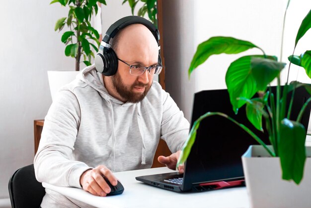 Homme adulte avec des lunettes et des écouteurs assis à une table travaillant en ligne sur un ordinateur portable à la maison