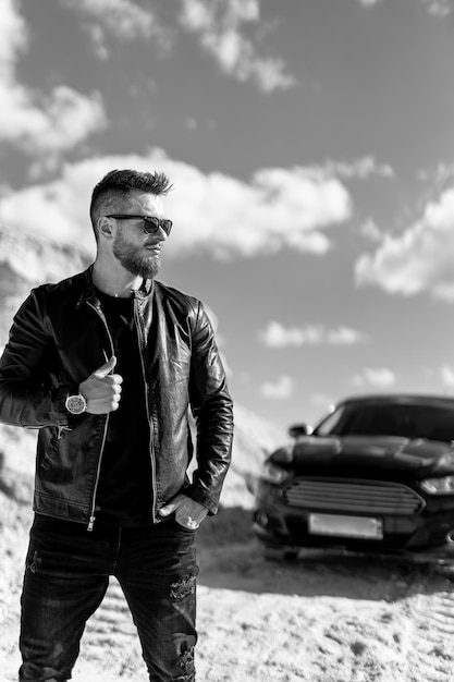 Homme adulte à la mode debout devant la voiture. Photographie monochrome. Homme tenant sa veste noire.