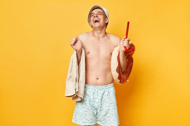 Homme adulte positif extrêmement heureux portant un maillot de bain short et panama tenant un anneau en caoutchouc de beignet et un sac isolé sur fond jaune se réjouissant reposant sur un complexe tropical