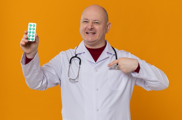 Homme adulte souriant en uniforme de médecin avec stéthoscope tenant un blister de médicament et pointant sur lui-même