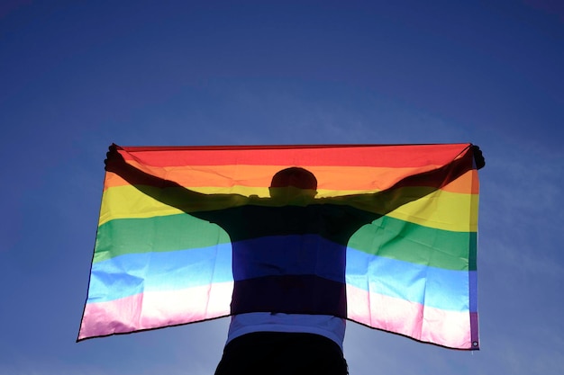 Un homme adulte en t-shirt blanc agite un drapeau arc-en-ciel avec un ciel bleu.