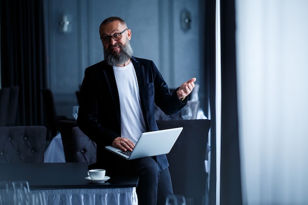 Un homme d'affaires adulte avec une barbe dans des lunettes est assis avec un ordinateur portable sur ses genoux et travaille. Le directeur établit un calendrier pour le flux de travail