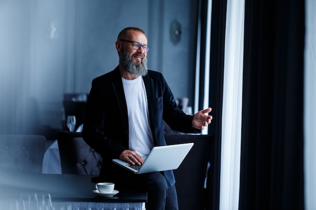 Un homme d'affaires adulte avec une barbe dans des lunettes est assis avec un ordinateur portable sur ses genoux et travaille. Le directeur établit un calendrier pour le flux de travail