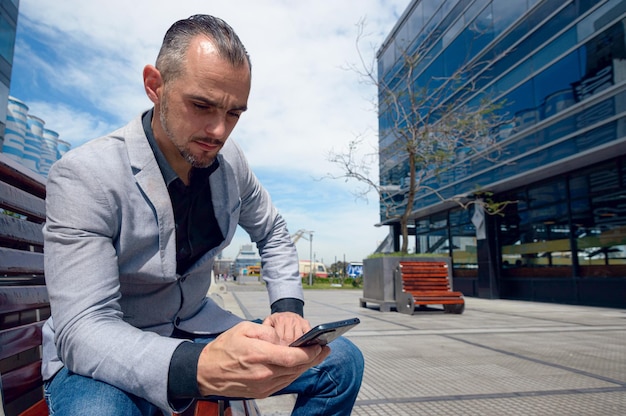 Homme d'affaires adulte caucasien français avec barbe portant des vêtements élégants assis à l'extérieur avec des bâtiments commerciaux autour en utilisant l'espace de copie de messages par téléphone