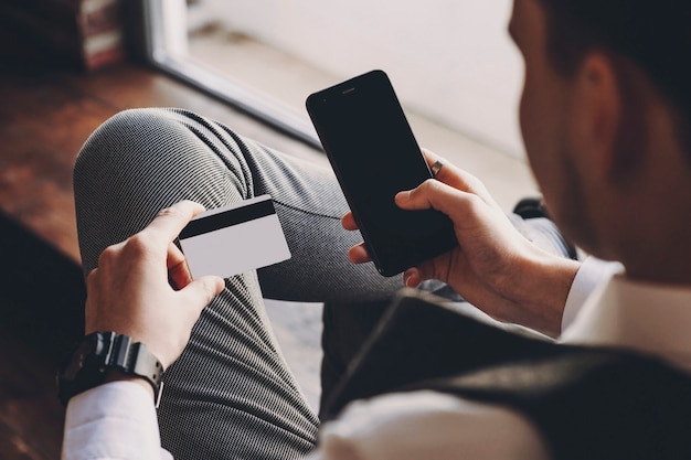 Homme d'affaires adulte confiant assis sur une chaise dans son entreprise près d'une fenêtre faisant une transaction d'argent avec carte sur le smartphone.