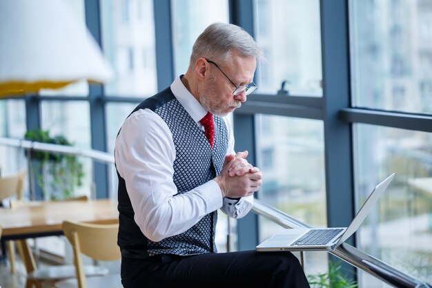 Homme d'affaires adulte, enseignant, mentor travaillant sur un nouveau projet. Assis près d'une grande fenêtre sur la table. Il regarde l'écran de l'ordinateur portable.