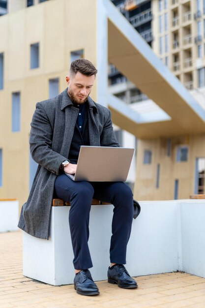 Homme d'affaires adulte en manteau gris assis avec un ordinateur portable Homme concentré travaillant dans la rue moderne avec une tasse de café