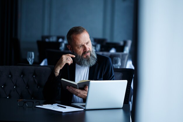 Un homme d'affaires adulte et prospère travaille sur un nouveau projet et examine les courbes de croissance dans un cahier. Assis à table près de la grande fenêtre. Regarde un cahier et sourit