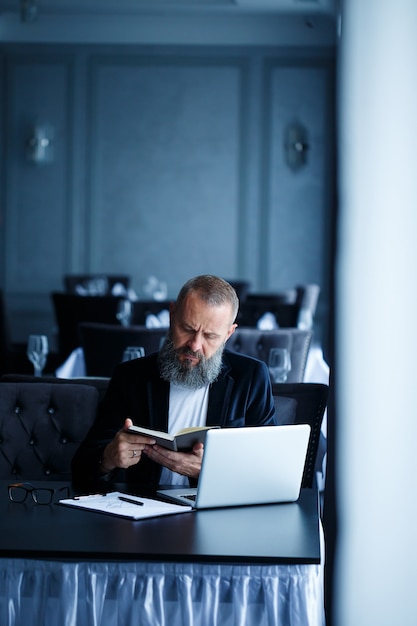 Un homme d'affaires adulte et prospère travaille sur un nouveau projet et examine les courbes de croissance dans un cahier. S'assoit à table et travaille. Regarde le cahier et sourit