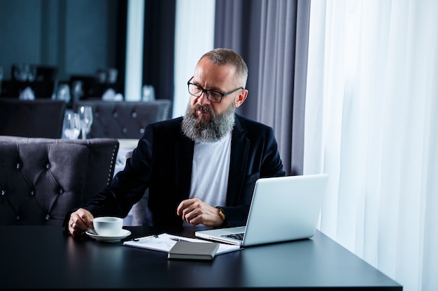 Un homme d'affaires adulte travaille sur un nouveau projet et examine les graphiques de croissance des actions. Assis à table près de la grande fenêtre. Regarde l'écran de l'ordinateur portable et boit du café.