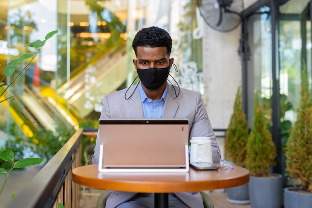 Homme d'affaires africain au café portant un masque facial tout en se distanciant socialement et en utilisant un ordinateur portable