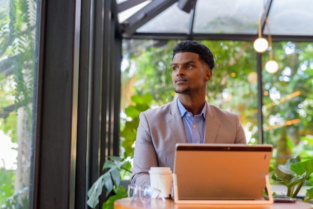 Homme d'affaires africain au café utilisant un ordinateur portable et pensant tout en regardant par la fenêtre