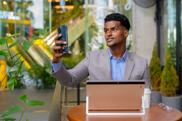 Homme d'affaires africain au café utilisant un ordinateur portable et prenant un selfie avec un téléphone portable