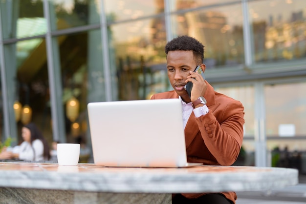 Homme d'affaires africain à l'extérieur dans un café utilisant un ordinateur portable tout en parlant sur un téléphone portable