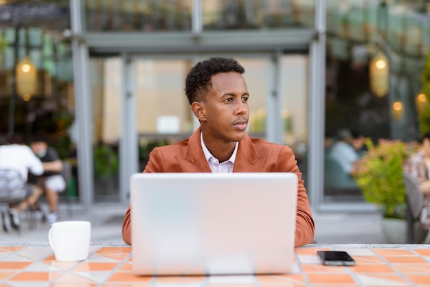 Homme d'affaires africain à l'extérieur dans un café utilisant un ordinateur portable tout en pensant