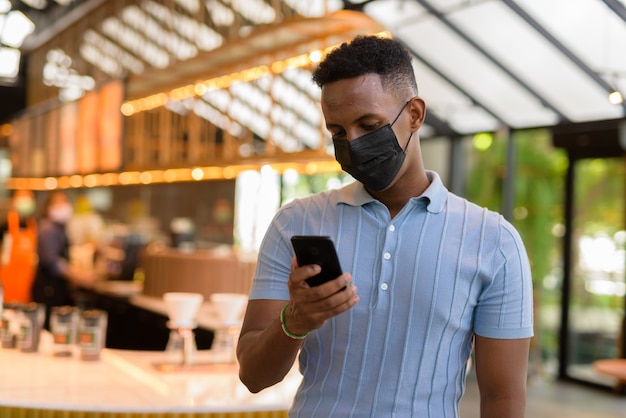 Homme d'affaires africain portant un masque facial dans un café-restaurant tout en se distanciant socialement et en utilisant un téléphone portable