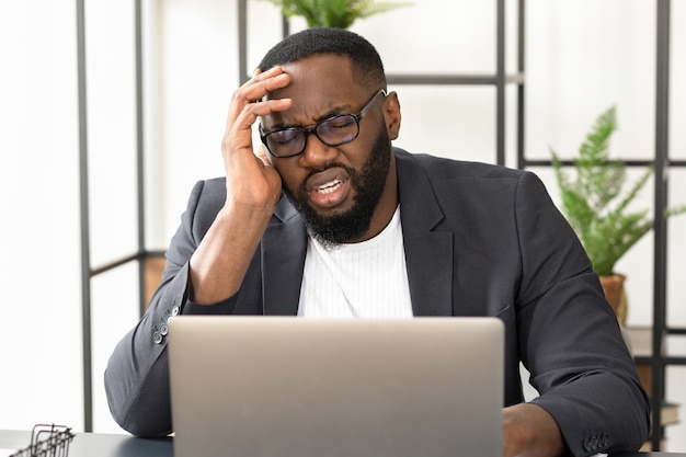 Homme d'affaires afro-américain fatigué ayant des maux de tête ou des migraines assis au bureau. Un jeune homme indépendant assis au bureau à domicile souffre de douleurs atroces aux tempes