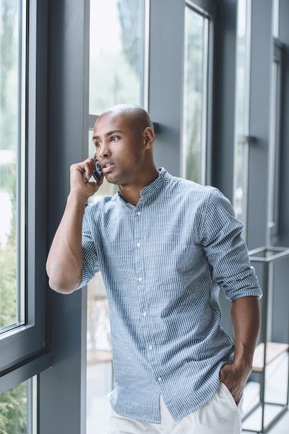 Homme d'affaires afro-américain réfléchi regardant par la fenêtre et parlant sur smartphone