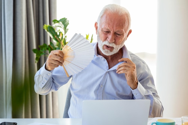 Un homme d'affaires âgé malade s'assoit sur un bureau pour travailler sur un ordinateur portable avec un ventilateur à main