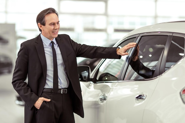 Homme d'affaires d'âge moyen en costume classique sourit en examinant une voiture dans un salon de l'automobile