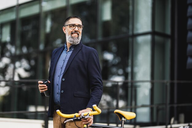 Homme d'affaires d'âge moyen prospère avec une tasse de café à emporter et avec un vélo à côté de lui, marche devant le quartier des bureaux.