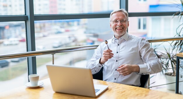 Homme d'affaires d'âge moyen souriant en chemise blanche avec un ordinateur portable. Homme assis près de la fenêtre travaillant avec des documents.