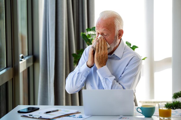 Un homme d'affaires âgé se sent malade avec le rhume et la fièvre au bureau malade avec la grippe assis à son bureau éternuer dans un mouchoir