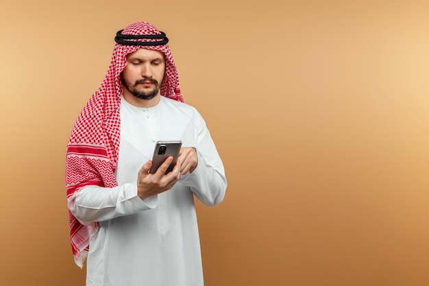 Homme d'affaires arabe en costume national tient un smartphone dans ses mains, mur beige.