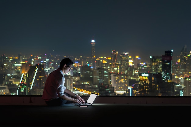 Homme d&#39;affaires asiatique assis et en utilisant l&#39;ordinateur portable sur le fond de paysage urbain pendant la nuit