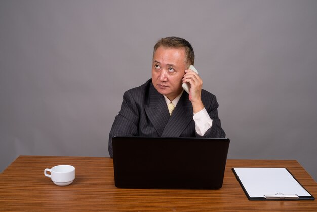 Homme d'affaires asiatique mature assis avec table en bois contre gris