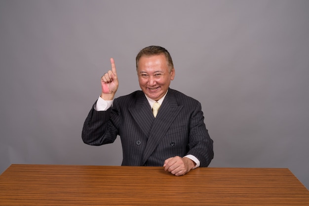 Homme d'affaires asiatique mature assis avec table en bois contre gris
