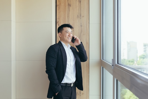 Homme d'affaires asiatique parlant au téléphone dans un centre de bureaux moderne partageant de bonnes nouvelles avec des compagnons