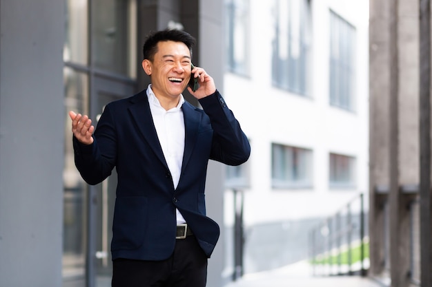 Un homme d'affaires asiatique parlant au téléphone, un sourire joyeux, rapporte de bonnes nouvelles près du centre de bureaux à l'extérieur