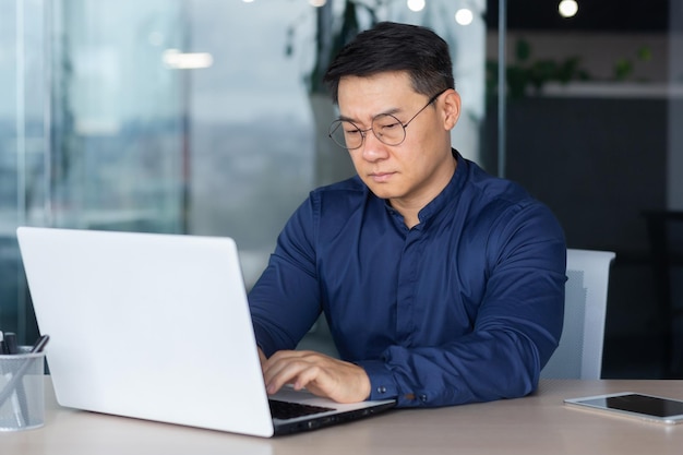 Homme d'affaires asiatique de pensée sérieuse travaillant à l'intérieur d'un bureau moderne homme mûr en chemise et lunettes