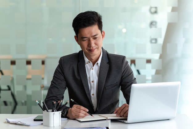 Un homme d'affaires asiatique porte un costume pour travailler au bureau en prenant des notes et un ordinateur portable.